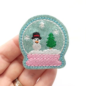 May include: A blue glitter snow globe with a white snowman and a green pine tree inside. The base of the snow globe is pink.