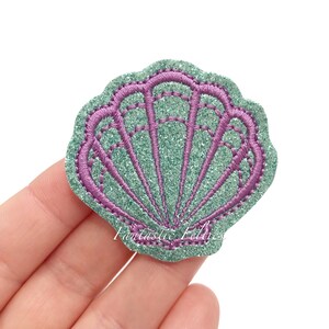 May include: A green glitter seashell with a purple outline. The seashell is a popular craft supply for making jewelry, hair accessories, and other projects.
