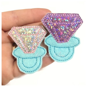 May include: Two felt diamond ring appliques with iridescent glitter fabric. The diamonds are pink and purple and are sitting on a light blue felt ring base.