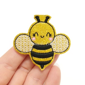 May include: A yellow and black glitter bee with white wings and a smiling face. The bee is embroidered with black thread and has a pink blush on its cheeks.