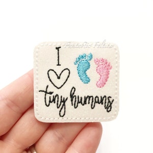 May include: White felt patch with black embroidery that reads "I heart tiny humans" with two embroidered footprints, one blue and one pink.