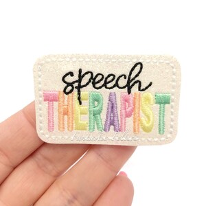 May include: A rectangular, glittery patch with the words "speech therapist" embroidered in black and pastel colors. The patch has a stitched border and the text "Fantastic Felties" is at the bottom.
