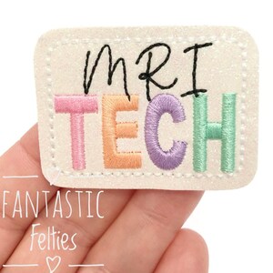 May include: A white felt patch with the words "MRI TECH" embroidered in colorful letters. The letters are pink, orange, purple, and green. The patch is surrounded by a white stitched border.