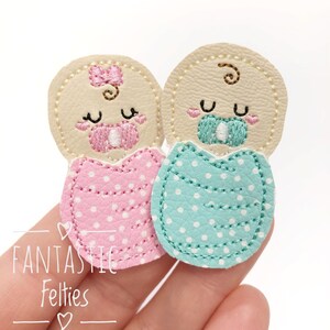 May include: Two felt baby appliques, one in pink and one in blue, with embroidered faces and pacifiers. The appliques are decorated with white polka dots.