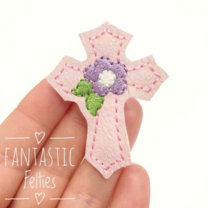 May include: A pink felt cross with a purple flower and green leaves embroidered on it. The cross is decorated with a pink stitch around the edges.