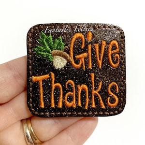 May include: A brown felt patch with glitter, featuring orange embroidery that reads "Give Thanks" and a green and white embroidered acorn.