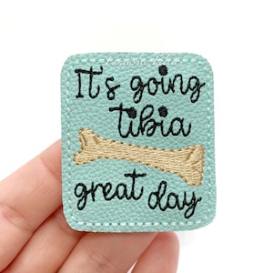 May include: A light blue rectangular patch with the text "It's going tibia great day" in black script. A tan bone graphic is embroidered below the word "tibia". The patch has a stitched border.