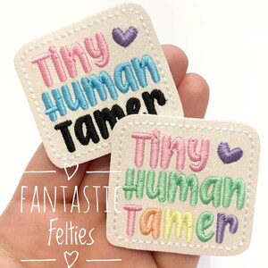 May include: Two white felt patches with the text "Tiny Human Tamer" in rainbow colors. The patches are embroidered with a small purple heart.