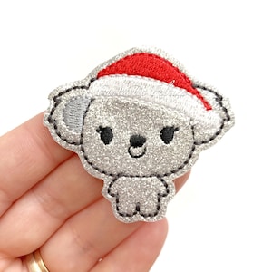 May include: A silver glitter koala wearing a red Santa hat. The koala has black eyes and a black nose. The koala is a Christmas-themed applique.