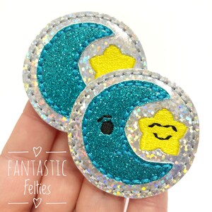 May include: Two turquoise blue felt moon and star appliques with silver glitter accents. The moon has a black eye and the star has a yellow center and a black smile.