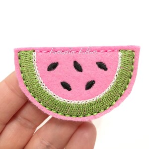 May include: A pink felt watermelon slice with green embroidered edges and black felt seeds.