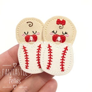 May include: Two felt baby appliques in a baseball-themed design. The appliques are white with red stitching and feature a baby face with a pacifier. One applique has a red bow on top of the head.
