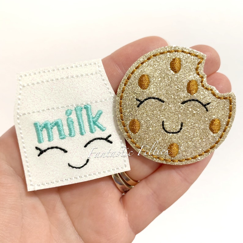 Puede incluir: Un conjunto de dos aplicaciones de fieltro, una una caja de leche con la palabra "leche" en letras turquesa y una cara sonriente, y la otra una galleta de purpurina dorada con chispas de chocolate y una cara sonriente.