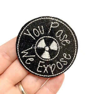 May include: Black and white embroidered patch with a glitter background. The patch is round and features a white radiation symbol with the text "You Pass We Expose" around it.