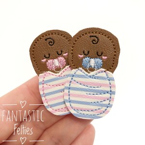 May include: Two felt baby appliques, one with a pink pacifier and pink and blue striped blanket, the other with a blue pacifier and blue and pink striped blanket.