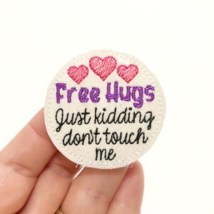 May include: White felt circle with pink hearts and the text "Free Hugs Just Kidding Don't Touch Me" in black and purple embroidery.