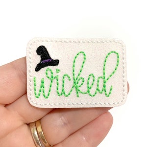 May include: A white felt patch with green embroidery that says "wicked" and a black witch hat with a purple band.