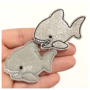 May include: Two felt shark appliques. One is gray and the other is silver glitter. Both have black embroidered eyes and mouths.