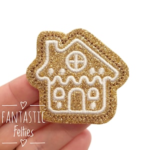 May include: Gold glitter gingerbread house applique with white stitching. The gingerbread house has a white window and a white icing design.