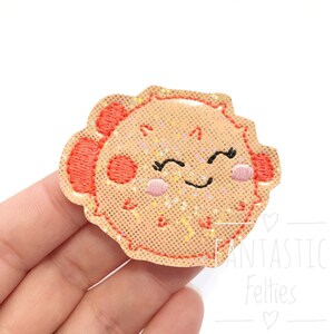 May include: A light pink and orange felt pufferfish with a smiling face and sparkly glitter. The pufferfish is a cute and whimsical design.