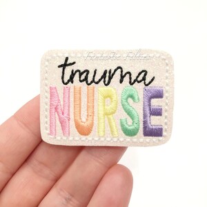 May include: A rectangular, off-white embroidered patch with the words "trauma NURSE" in black and rainbow-colored thread. The patch has a glittery texture and a stitched border. The text "Fantastic Felties" is written in cursive.
