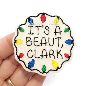 May include: White felt Christmas ornament with a string of colorful lights around the edge. The ornament says "It's a beaut, Clark!"
