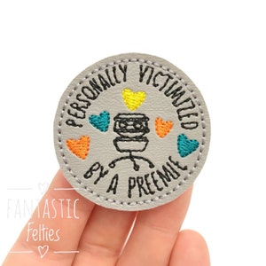 May include: A gray felt patch with black embroidery that reads "Personally victimized by a preemie" with a cartoon image of a baby monitor and colorful hearts.