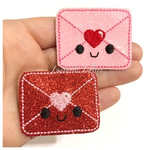 May include: Two felt envelope patches with a cute cartoon face and a heart. The envelope on the left is red with a pink heart. The envelope on the right is pink with a red heart.  Fantastic Felties.