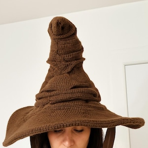 Könnte beinhalten: Ein brauner, gestrickter Sortierhut mit breiter Krempe und hoher, spitzer Krone. Der Hut hat ein strukturiertes Aussehen und ist mit mehreren Falten und Falten versehen. Der Hut ist ein Kostümzubehör.