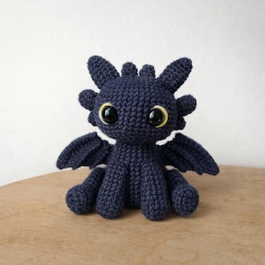 May include: A dark blue crochet dragon toy with large, round eyes and small wings. The dragon has a friendly expression and is sitting on a wooden surface. The dragon's eyes have yellow accents.