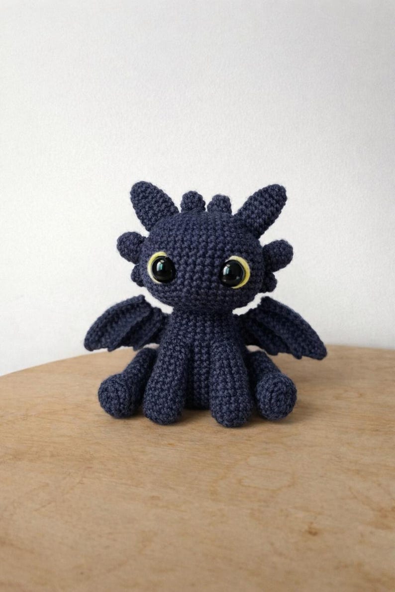 May include: A dark navy blue crochet plush toy shaped like a dragon with large, glossy black eyes surrounded by thin yellow rings. The creature features pointed ears, small head spikes, and two small wings extending from its sides while sitting upright.
