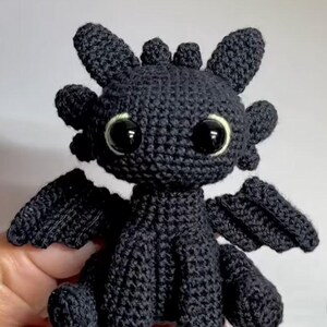 May include: A black crocheted dragon plush toy featuring large, glossy black eyes with thin light-colored rims, pointed ears, small horns on the head, and textured wings. The creature is crafted with visible yarn stitches in a seated position.