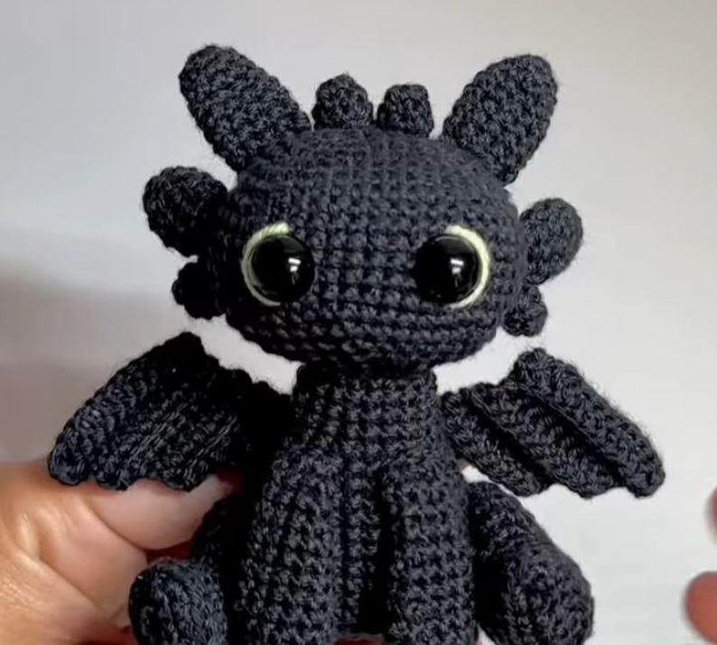 May include: A black crocheted dragon plush toy featuring large, glossy black eyes with thin light-colored rims, pointed ears, small horns on the head, and textured wings. The creature is crafted with visible yarn stitches in a seated position.