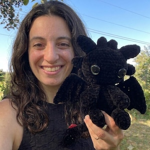 May include: A black crocheted dragon plush toy featuring large, glossy black eyes, pointed ears, small wings, and a tail with a small red accent at the tip. The creature is crafted from a soft, textured yarn and displays a detailed, rounded head.