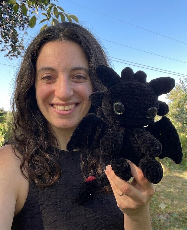 May include: A black crocheted dragon plush toy featuring large, glossy black eyes, pointed ears, small wings, and a tail with a small red accent at the tip. The creature is crafted from a soft, textured yarn and displays a detailed, rounded head.