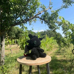May include: A black crocheted dragon plush toy with large, dark, reflective eyes, small wings, and a red tail tip. The plush features a rounded head with small ear-like protrusions and sits on a round wooden stool. The text KROKMOU is visible.