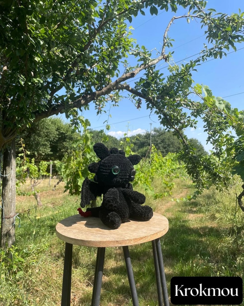 May include: A black crocheted dragon plush toy with large, dark, reflective eyes, small wings, and a red tail tip. The plush features a rounded head with small ear-like protrusions and sits on a round wooden stool. The text KROKMOU is visible.