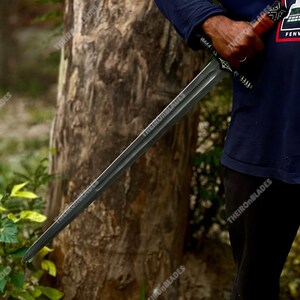 May include: A long, dark gray sword with a detailed handle and a straight blade. The sword is held in front of a tree, with the text "THEIRONBLADES" visible on the blade and handle.