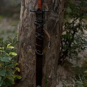 May include: A black sword with a brown handle and a leather-wrapped grip is leaning against a tree. The sword has a silver blade and a leather sheath. The handle has a decorative design.