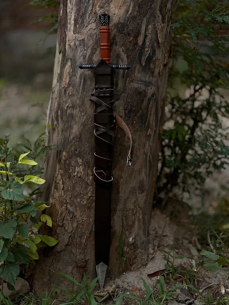 May include: A black sword with a brown handle and a leather-wrapped grip is leaning against a tree. The sword has a silver blade and a leather sheath. The handle has a decorative design.
