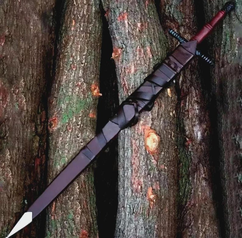 May include: A long, dark burgundy sword with a white tip, black crossguard, and a wrapped black handle. The sword is displayed against a backdrop of textured tree trunks, showcasing its craftsmanship and design.