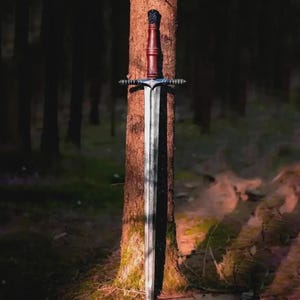 May include: A long, silver sword with a brown handle and a decorative crossguard is leaning against a tree in a forest. The sword's blade is long and sharp, and the handle is a dark brown color. The crossguard is silver with decorative elements.