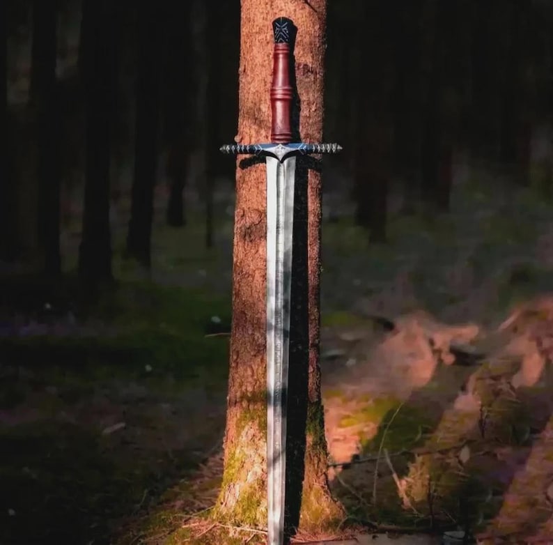 May include: A long, silver sword with a brown handle and a decorative crossguard is leaning against a tree in a forest. The sword's blade is long and sharp, and the handle is a dark brown color. The crossguard is silver with decorative elements.