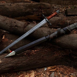 May include: A long, silver sword with a brown leather-wrapped handle and a black scabbard. The sword rests on a pile of wooden logs, with the scabbard beside it. The handle has a decorative design.
