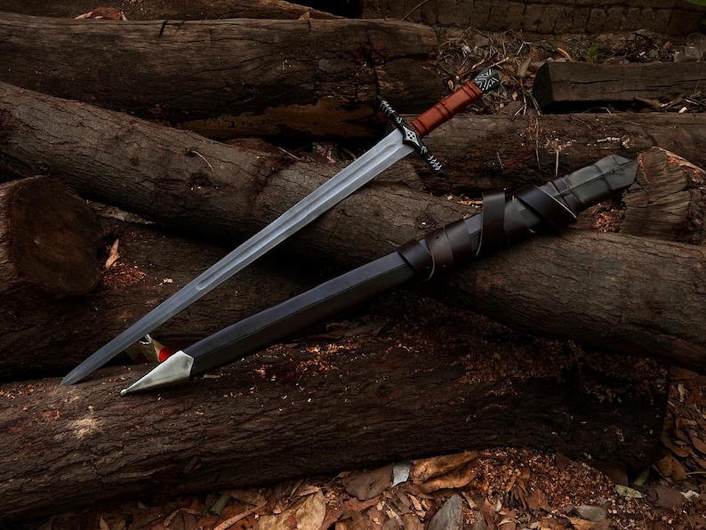 May include: A long, silver sword with a brown leather-wrapped handle and a black scabbard. The sword rests on a pile of wooden logs, with the scabbard beside it. The handle has a decorative design.