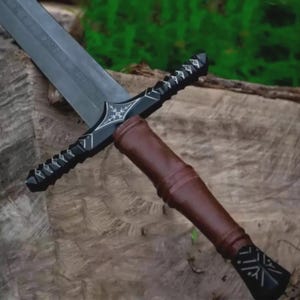 May include: A medieval-style sword with a dark blade and a black hilt featuring decorative silver accents. The handle is wrapped in brown leather. The sword is resting on a wooden surface.