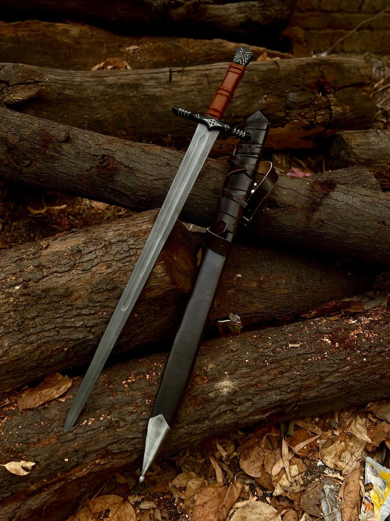 May include: A long, silver sword with a brown leather-wrapped handle and a black crossguard. The sword is sheathed in a black leather scabbard with a silver tip. The sword and scabbard are resting on a pile of wooden logs.