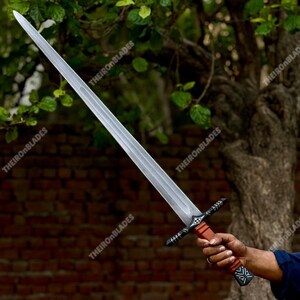 May include: A long, silver sword with a double-edged blade. The hilt is brown and black, with decorative patterns. The sword is held against a backdrop of green foliage and a brick wall.