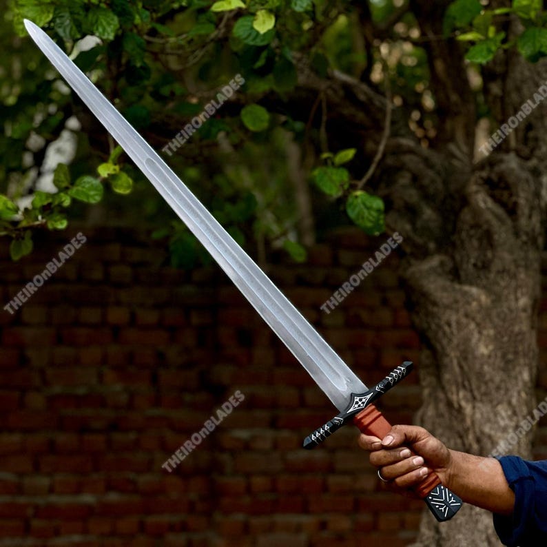 May include: A long, silver sword with a double-edged blade. The hilt is brown and black, with decorative patterns. The sword is held against a backdrop of green foliage and a brick wall.