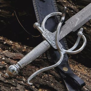 May include: A silver rapier sword with a decorative handle and guard, resting in a black leather sheath. The blade has a patterned design. The sword is displayed on a textured, brown surface.